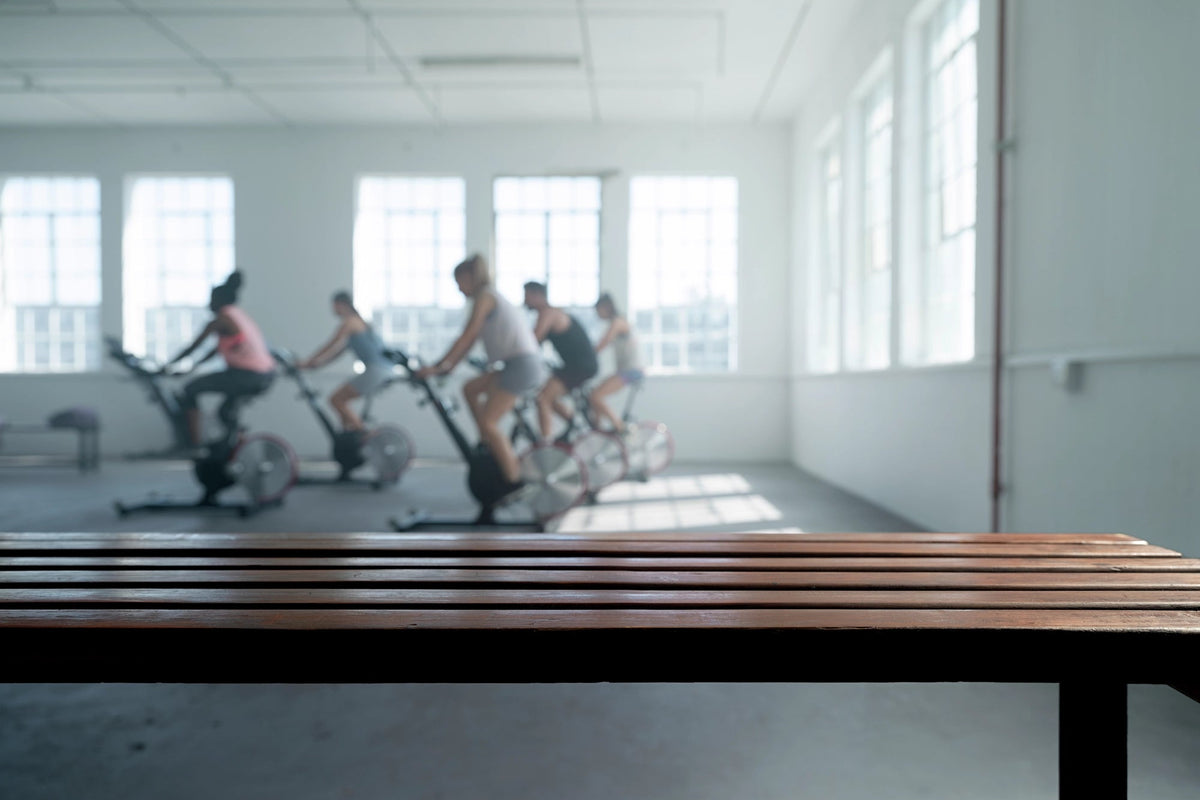 People exercising on stationary bikes in a gym with a wooden bench in the foreground.